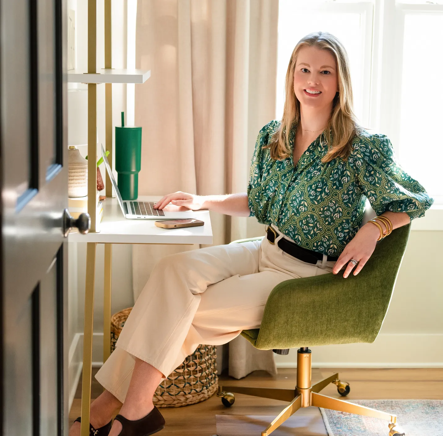 about-1 Woman in a green patterned blouse sits at a desk with a laptop, smiling at the camera in a bright room.