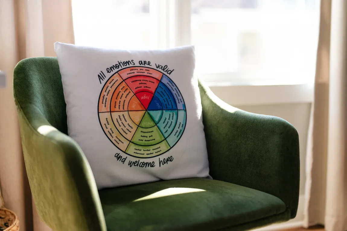 about-3 A green chair holds a pillow displaying a colorful emotion wheel and the phrase "All emotions are valid and welcome here.