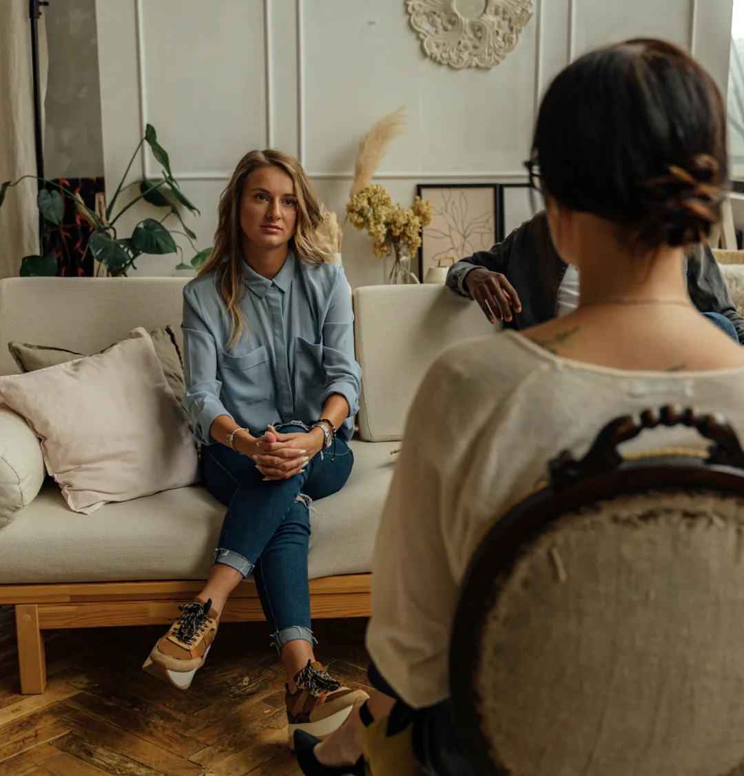Two women sit facing each other in conversation on a sofa and chair in a cozy, well-decorated room.