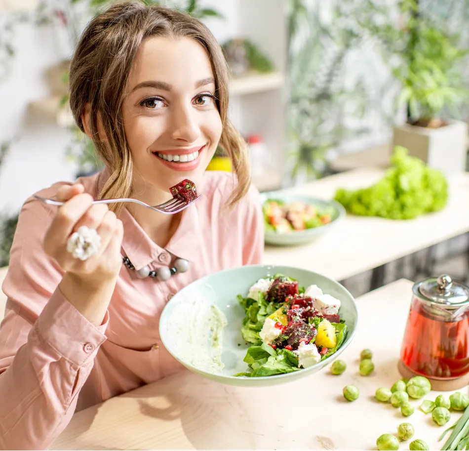 Smiling woman in a pink shirt eating a fresh salad at a bright, plant-filled kitchen table.