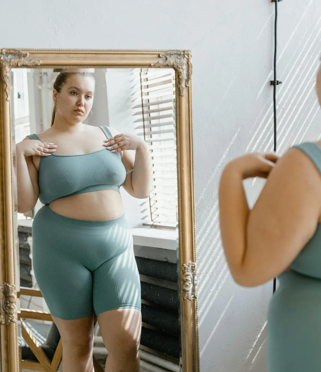 body-image-1 Woman in teal activewear looks at her reflection in a full-length mirror in a bright room.