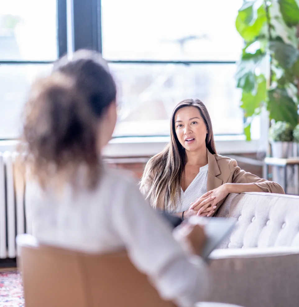Two women sit on couches talking in a bright, modern office with plants and large windows.
