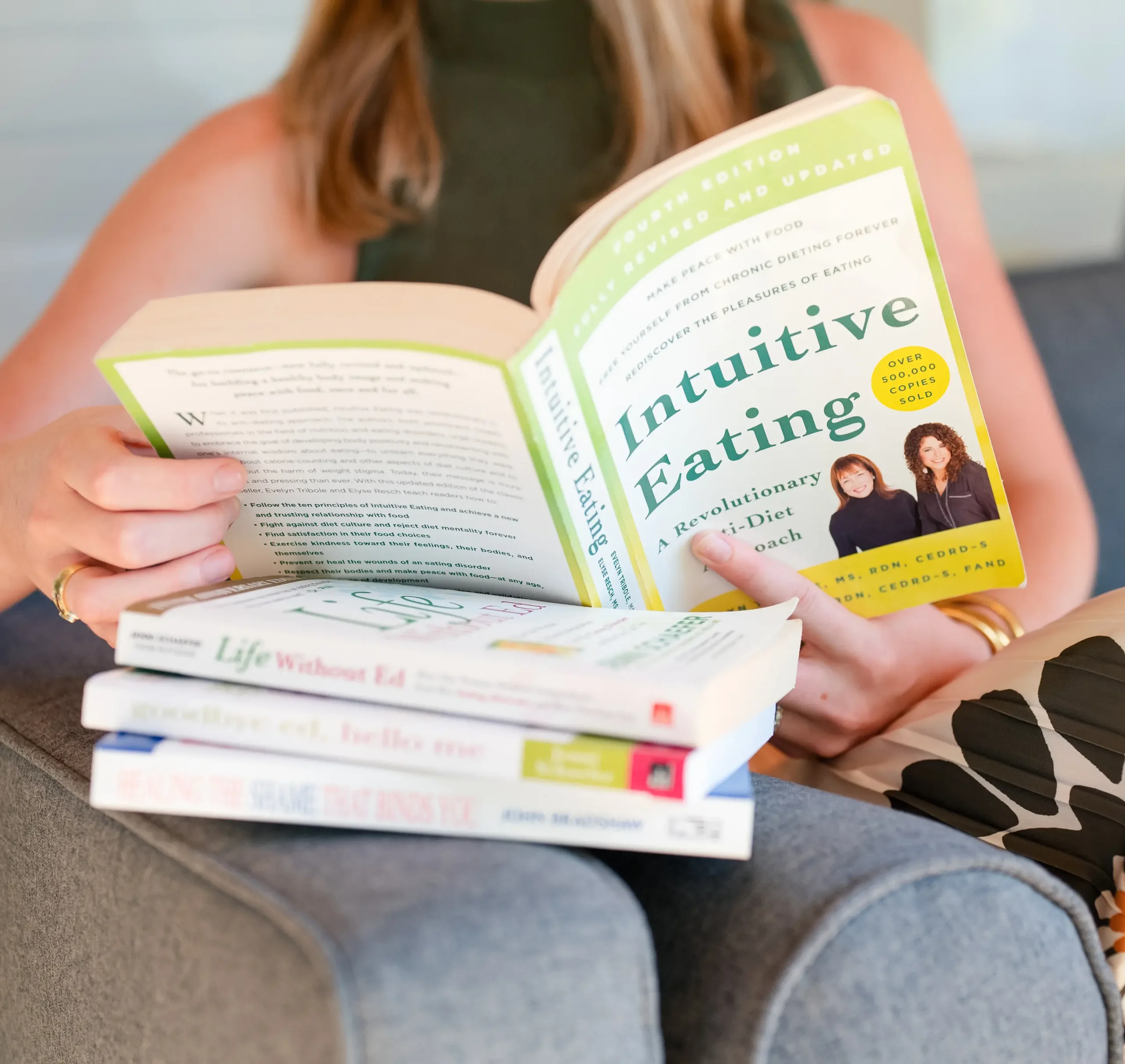 ed-1 A woman sits on a couch reading "Intuitive Eating," with three other books stacked on her lap.