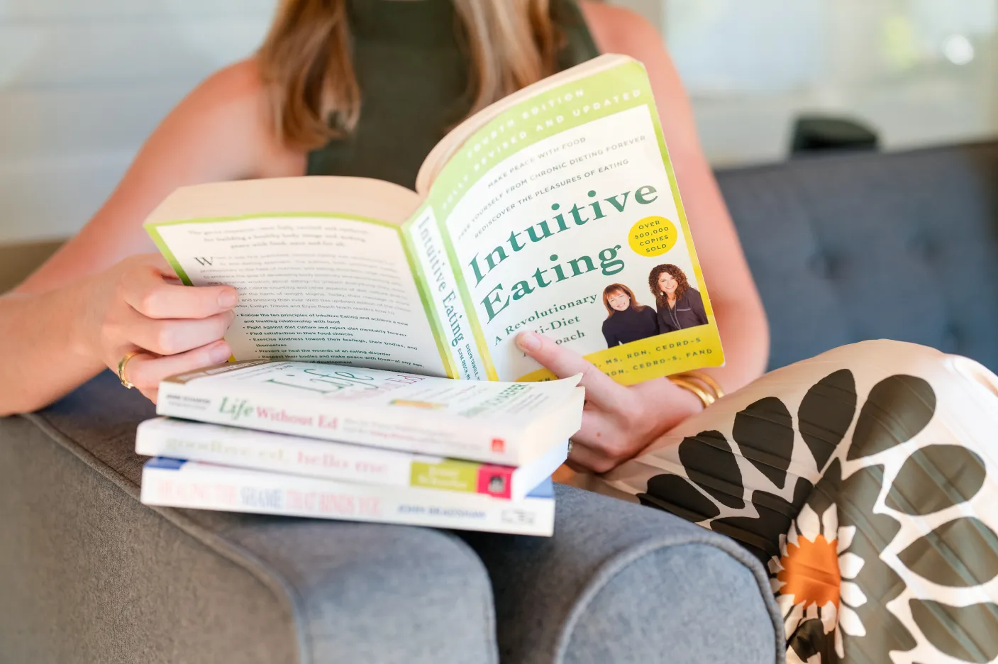 exp-1 A woman sits on a couch reading "Intuitive Eating," with three other books stacked beside her.