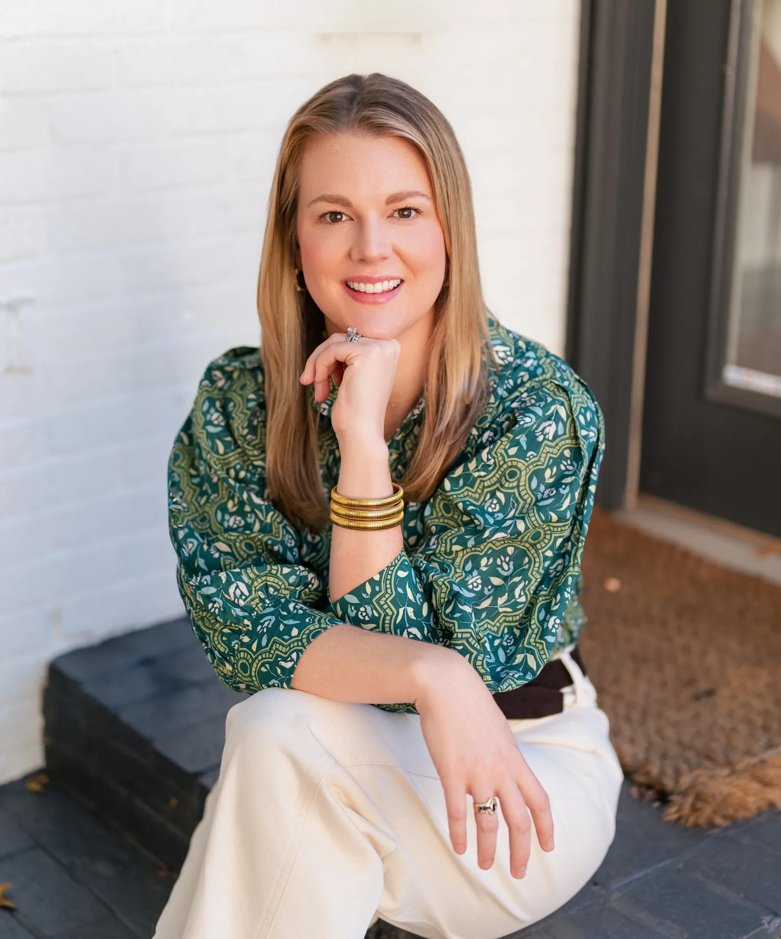 hero-1 A woman with blonde hair sits on a step, smiling, wearing a green patterned top and white pants.