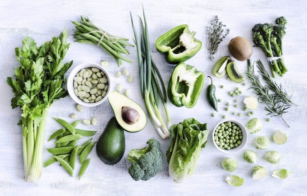 Assorted green vegetables and herbs arranged on a light surface, including avocado, celery, and green beans.