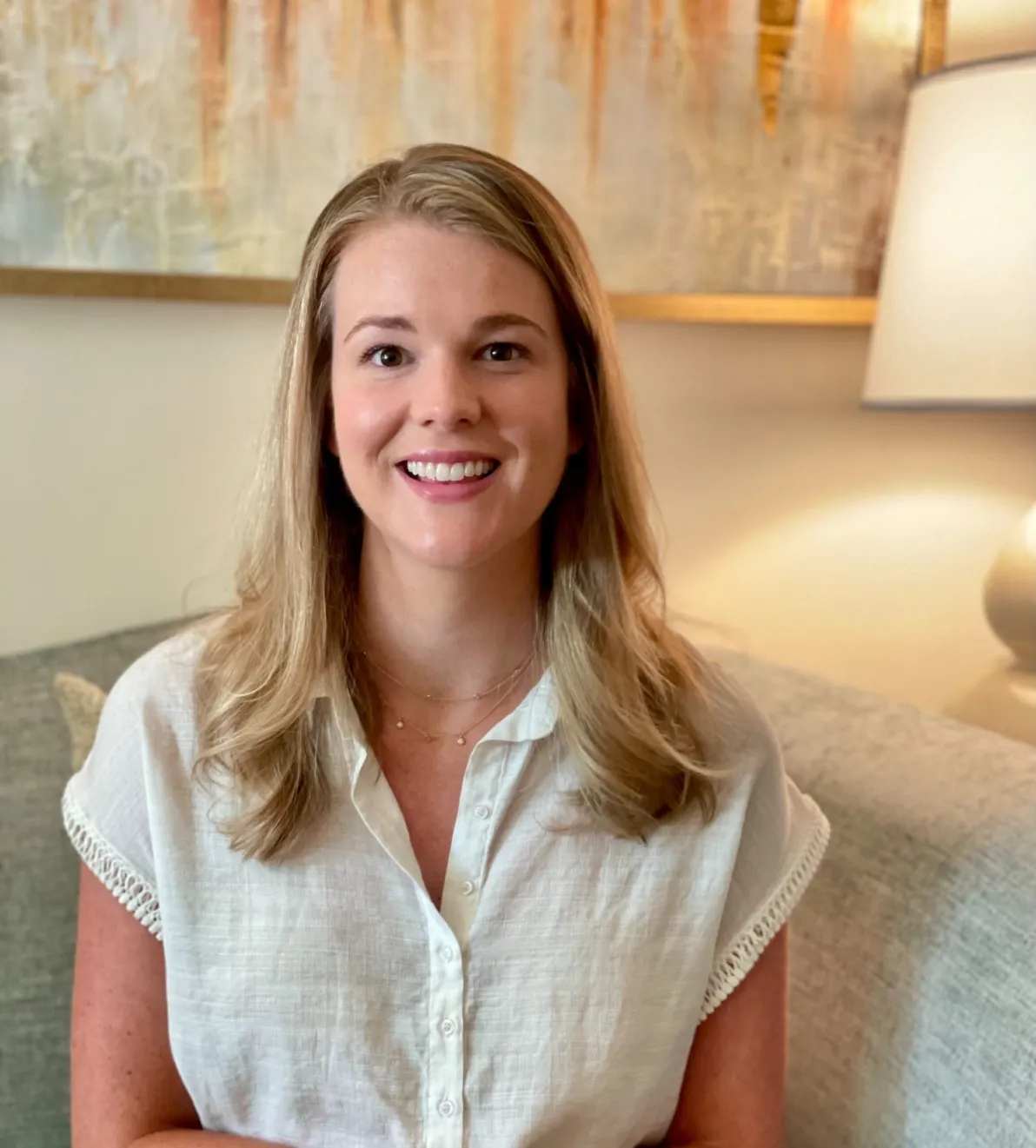 sarah-1 A smiling woman with blonde hair sits on a couch in a well-lit room with a lamp and abstract art behind her.
