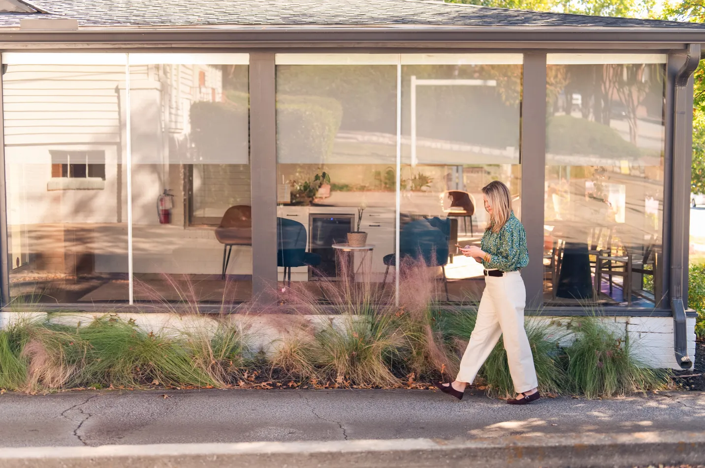 Woman walking outside a modern building with large windows, holding a phone and a drink.