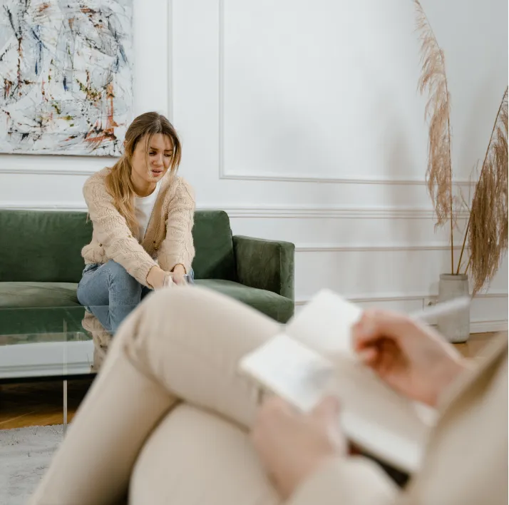 A woman sits on a couch looking pensive while another person takes notes in a notebook.