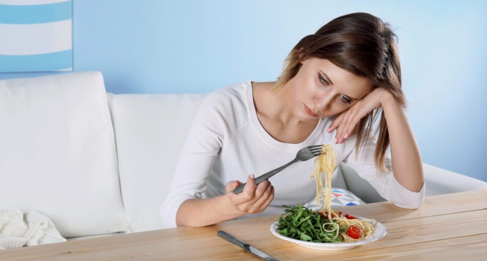 A woman looks bored while eating pasta and salad at a table.