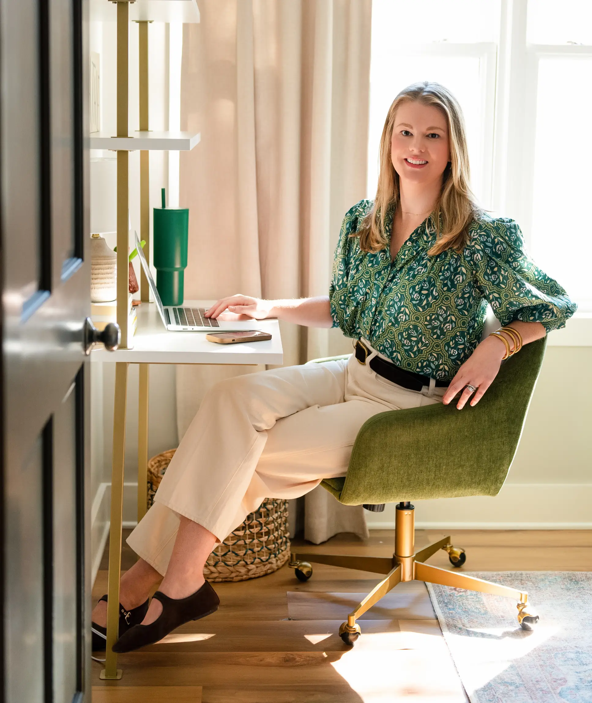 telehealth-2 Woman sitting at a desk with a laptop, smiling, in a bright home office with neutral and green decor.