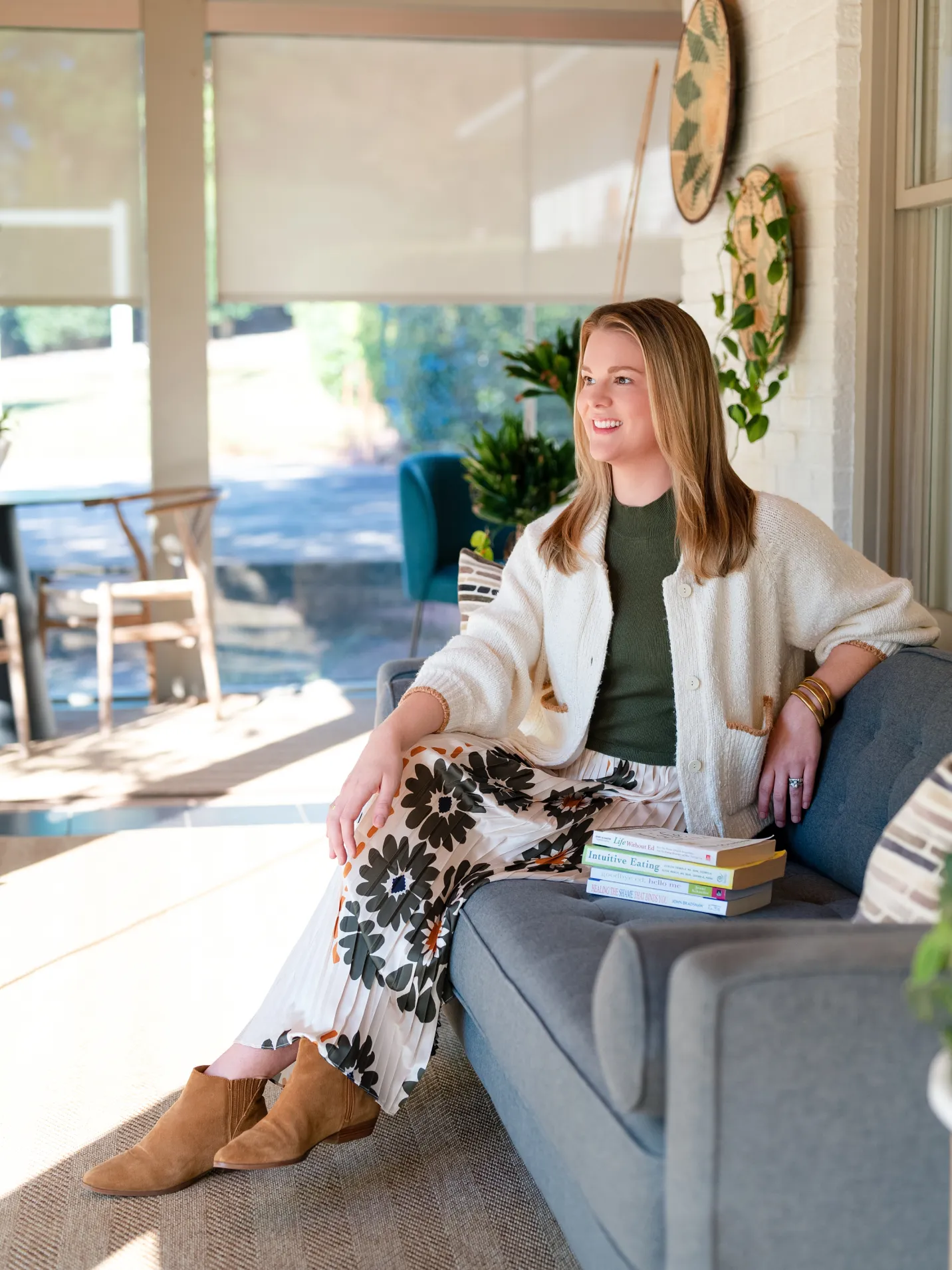 Woman sitting on a gray couch with books, wearing a floral skirt and cardigan, smiling in a bright room.