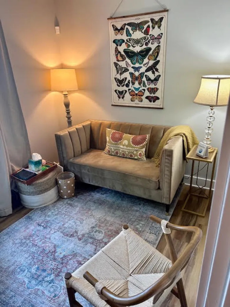 Cozy living room with a tan sofa, butterfly wall art, two lamps, a rug, and a woven chair.