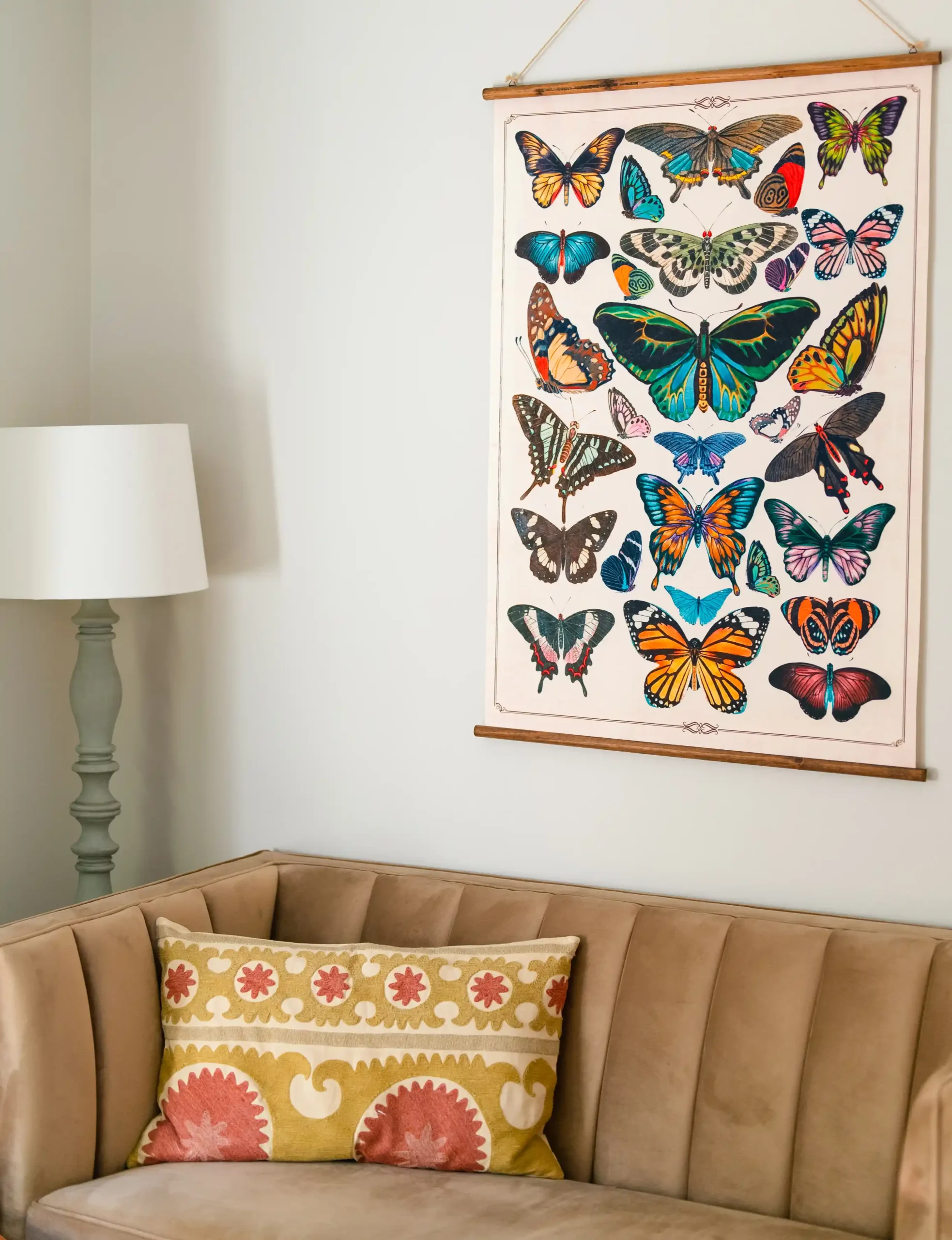 Wall art of colorful butterflies above a tan sofa with a patterned pillow and a floor lamp beside it.