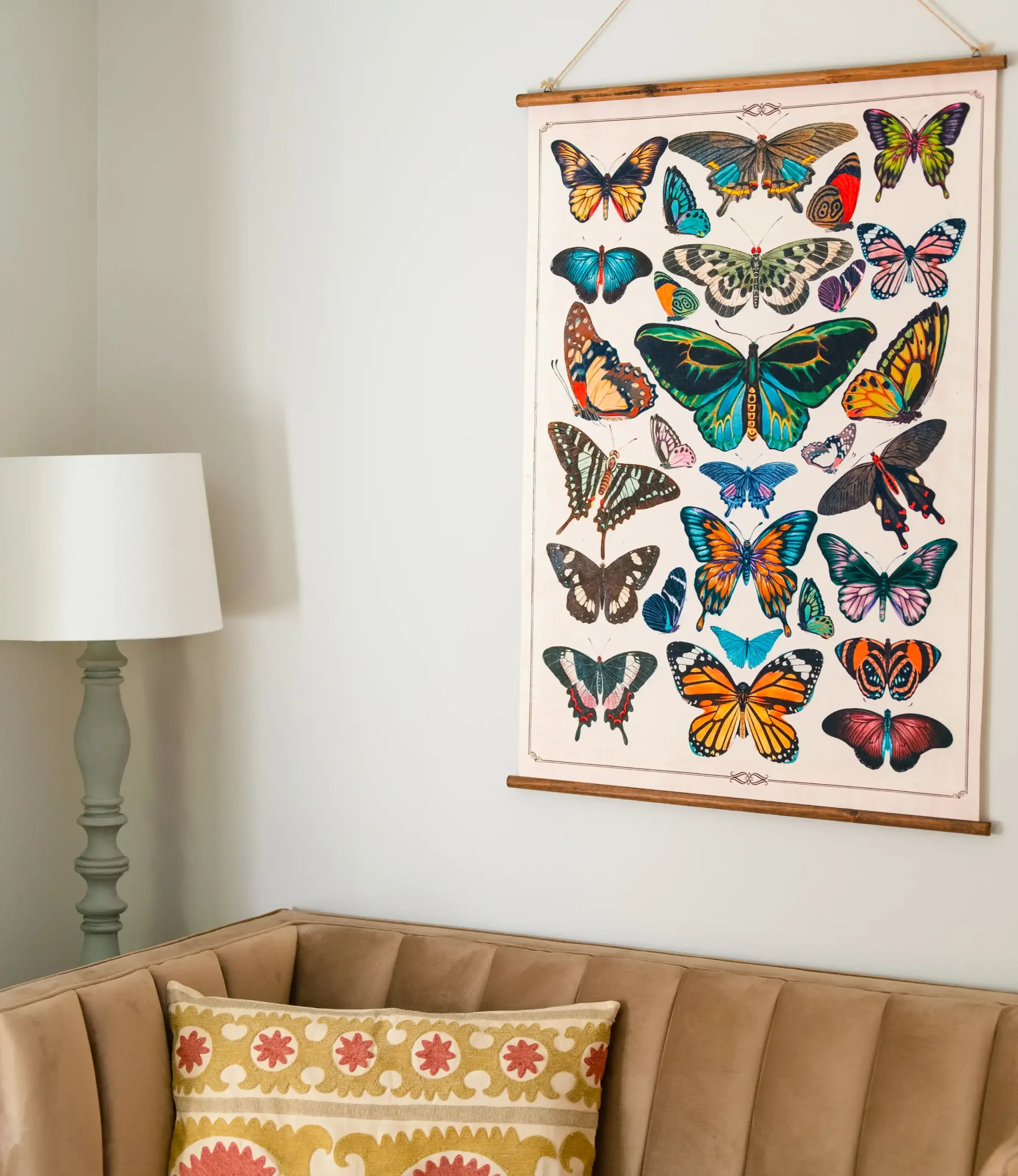 why-choose-4 Colorful butterfly print hanging above a tan couch with a patterned pillow and a green lamp beside it.